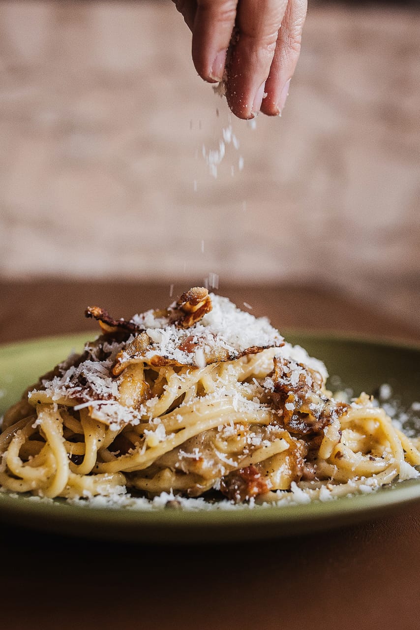 Carbonara sendo finalizada à mão com queijo, na cozinha da trattoria